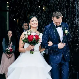 Uśmiechnięta panna młoda w białej sukni i pan młody w granatowym smokingu wychodzą z kościoła w deszczu, trzymając się za ręce. W tle widać gości weselnych.