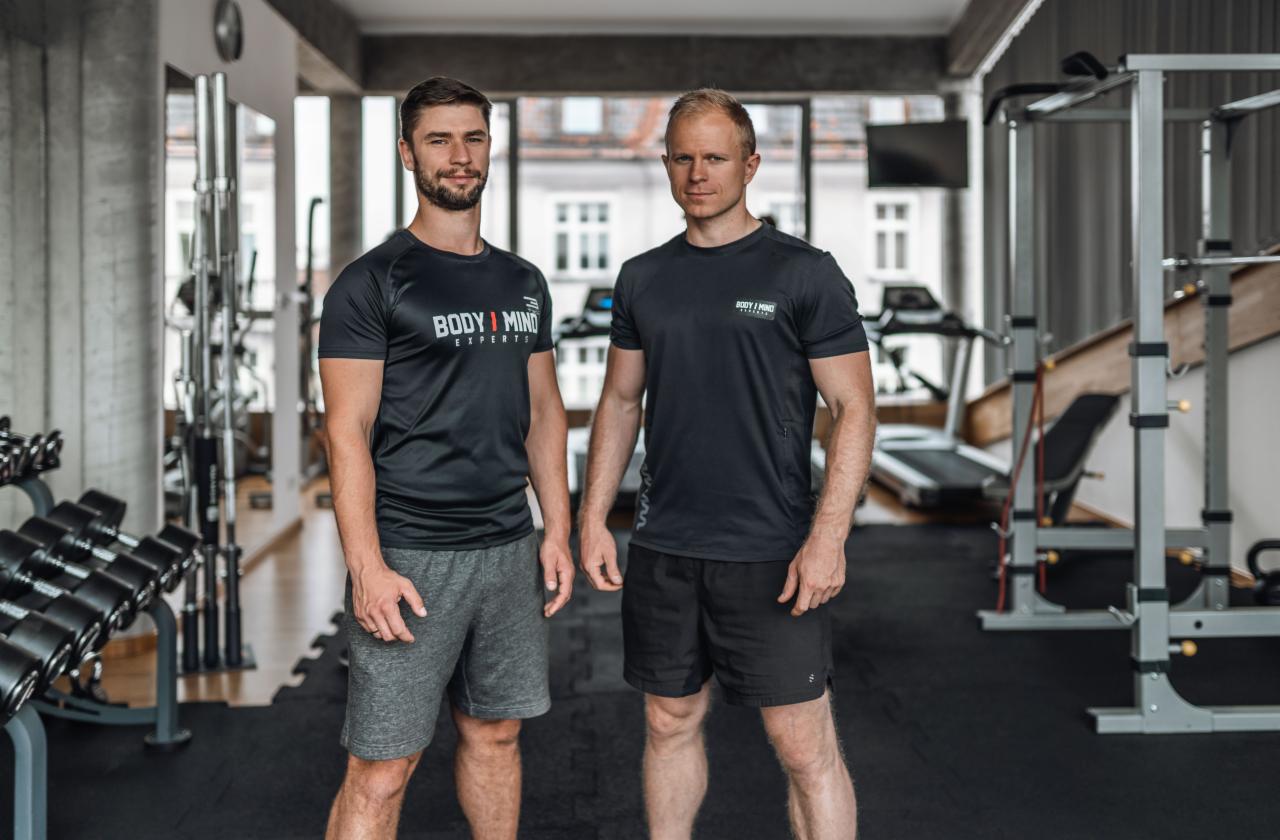 Dwóch mężczyzn w sportowych koszulkach z logo w siłowni, obok regału z hantlami i sprzętu do ćwiczeń.