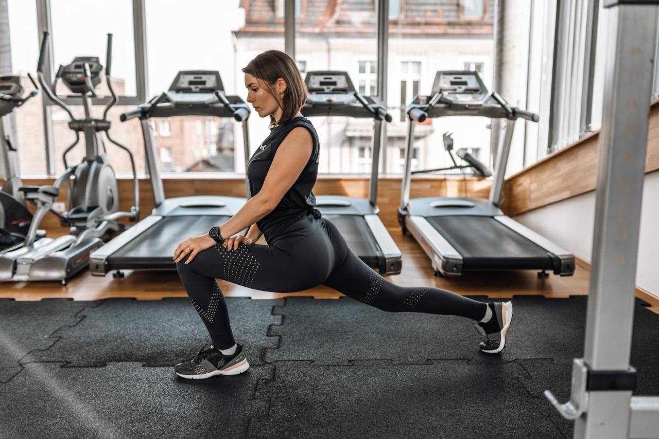 Kobieta w czarnym stroju sportowym rozciąga się, wykonując wypad w przód na tle sprzętu do ćwiczeń w siłowni, z widocznym zegarkiem sportowym na nadgarstku.