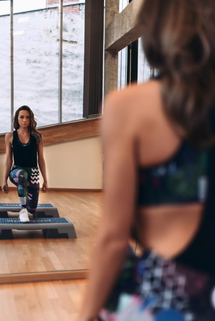 Kobieta w sportowym stroju wykonuje wypad na stepach w siłowni, widoczna w lustrze, z napisem na szybie w tle.