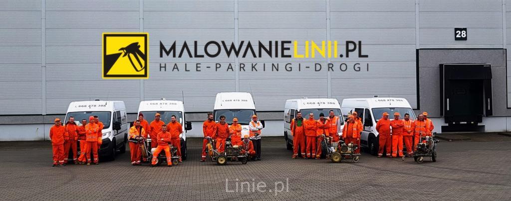 Grupa pracowników w pomarańczowych kombinezonach i z maszynami do malowania linii stoi przed budynkiem magazynowym z logo firmy MalowanieLinii.pl i numerem 28 nad rampą załadunkową.