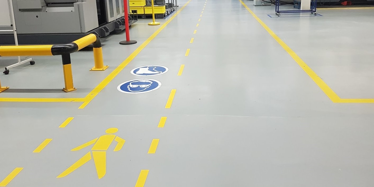 Oznakowanie poziome w hali produkcyjnej: szara posadzka z żółtymi liniami wyznaczającymi ciągi komunikacyjne i strefy bezpieczeństwa, w tym piktogramy nakazu stosowania ochrony oczu i obuwia...