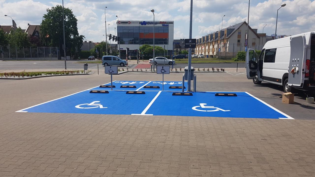 Niebieskie, świeżo odmalowane miejsca parkingowe dla osób niepełnosprawnych z białymi symbolami, białe linie oddzielające miejsca, biały dostawczy samochód z otwartymi drzwiami w tle, widoczne...