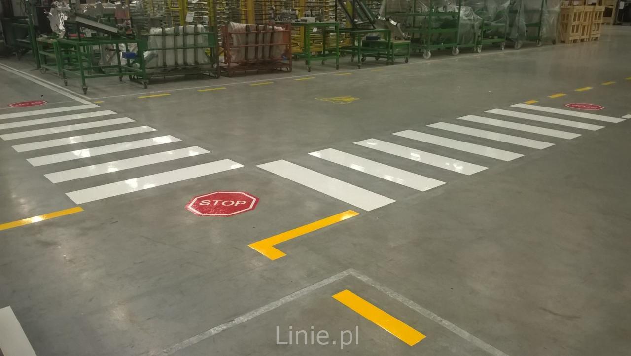 Oznakowanie poziome w hali produkcyjnej: białe pasy przejść dla pieszych, żółte linie i znaki STOP na szarym, betonowym podłożu.