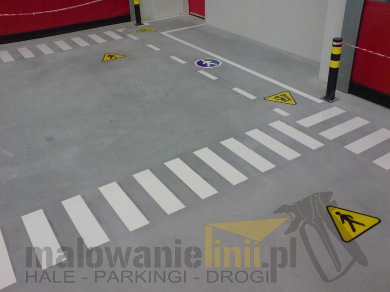 Oznakowanie poziome w hali: białe pasy przejścia dla pieszych, żółte trójkątne znaki ostrzegawcze i okrągły znak informacyjny na szarym betonie. Widoczny słup zabezpieczający w czarno-żółte pasy...
