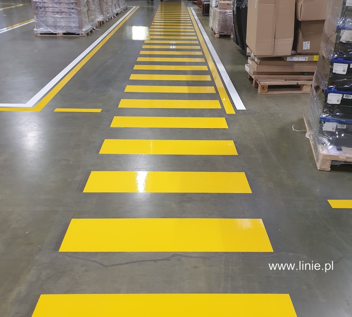 Żółte pasy na szarym betonowym podłożu w hali magazynowej, wyznaczające ciąg komunikacyjny, z widocznymi paletami i kartonami w tle.