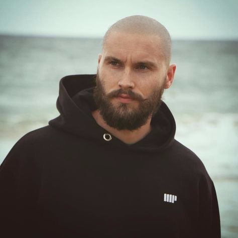 Mężczyzna z mikropigmentacją skóry głowy, brodą i wąsami, w czarnej bluzie z kapturem na tle morza.