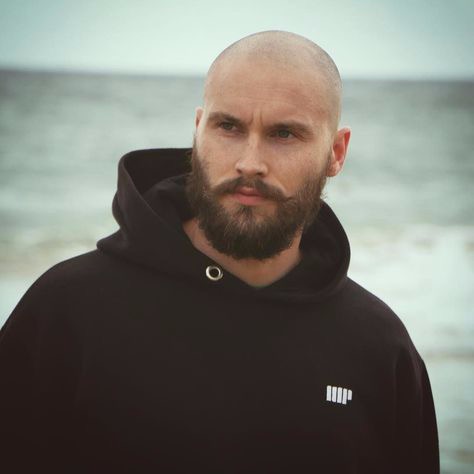 Portret łysiejącego mężczyzny z brodą i wąsami w czarnej bluzie z kapturem na tle morza.