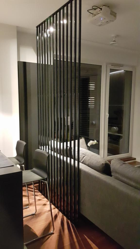 Metalowa ścianka działowa z pionowych prętów w nowoczesnym salonie, rzucająca cienie na drewnianą podłogę, z widocznym projektorem sufitowym.