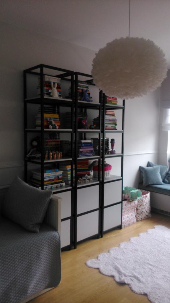 Metalowa konstrukcja regału z białymi półkami, wypełniona książkami i dekoracjami, pod puchową lampą w pokoju dziecięcym.