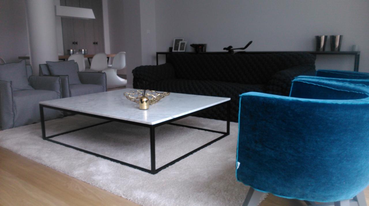 Nowoczesny salon z marmurowym stolikiem kawowym na czarnym stelażu, turkusowym fotelem i czarną sofą w tle, oświetlony naturalnym światłem.