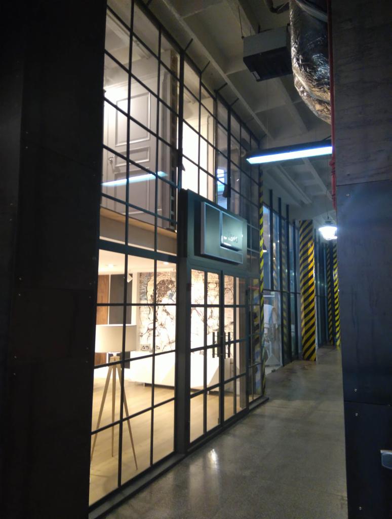 Wnętrze budynku z przeszkloną ścianą z czarnymi szprosami, widoczny fragment pomieszczenia z lampą na trójnogu i dekoracyjną tapetą.