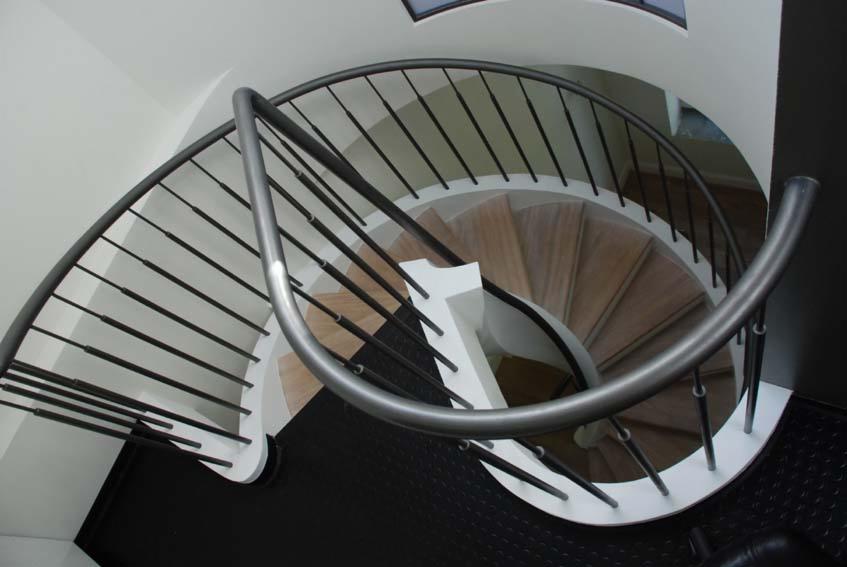 Spiralne schody z drewnianymi stopniami i metalową balustradą, widok z góry, czarna podłoga z fakturą w tle.