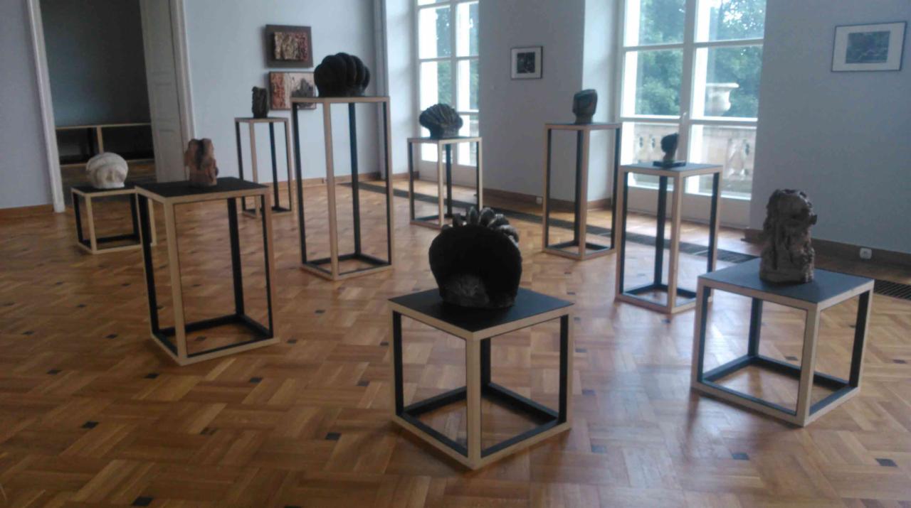 Wystawa rzeźb w jasnym pomieszczeniu z parkietem w jodełkę, eksponaty na minimalistycznych, kwadratowych postumentach z jasnego drewna i czarnymi krawędziami.