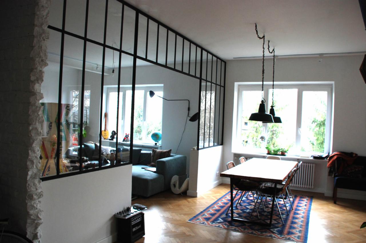 Nowoczesna, czarna ścianka działowa z metalowymi profilami i przeszkleniami oddzielająca salon od jadalni, widoczna z bliska, z fragmentem surowej ściany z cegły po lewej stronie.