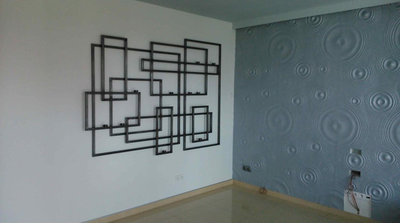 Metalowa dekoracja ścienna z połączonych kwadratów o surowym wykończeniu, na tle białej ściany i ściany z dekoracyjną tapetą w okręgi.