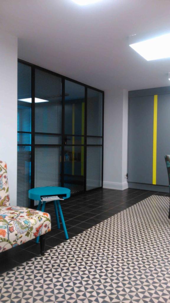 Nowoczesne biuro z metalową ścianką działową z żaluzjami, turkusowym stolikiem i wzorzystą podłogą, akcentowane żółtymi paskami na drzwiach.