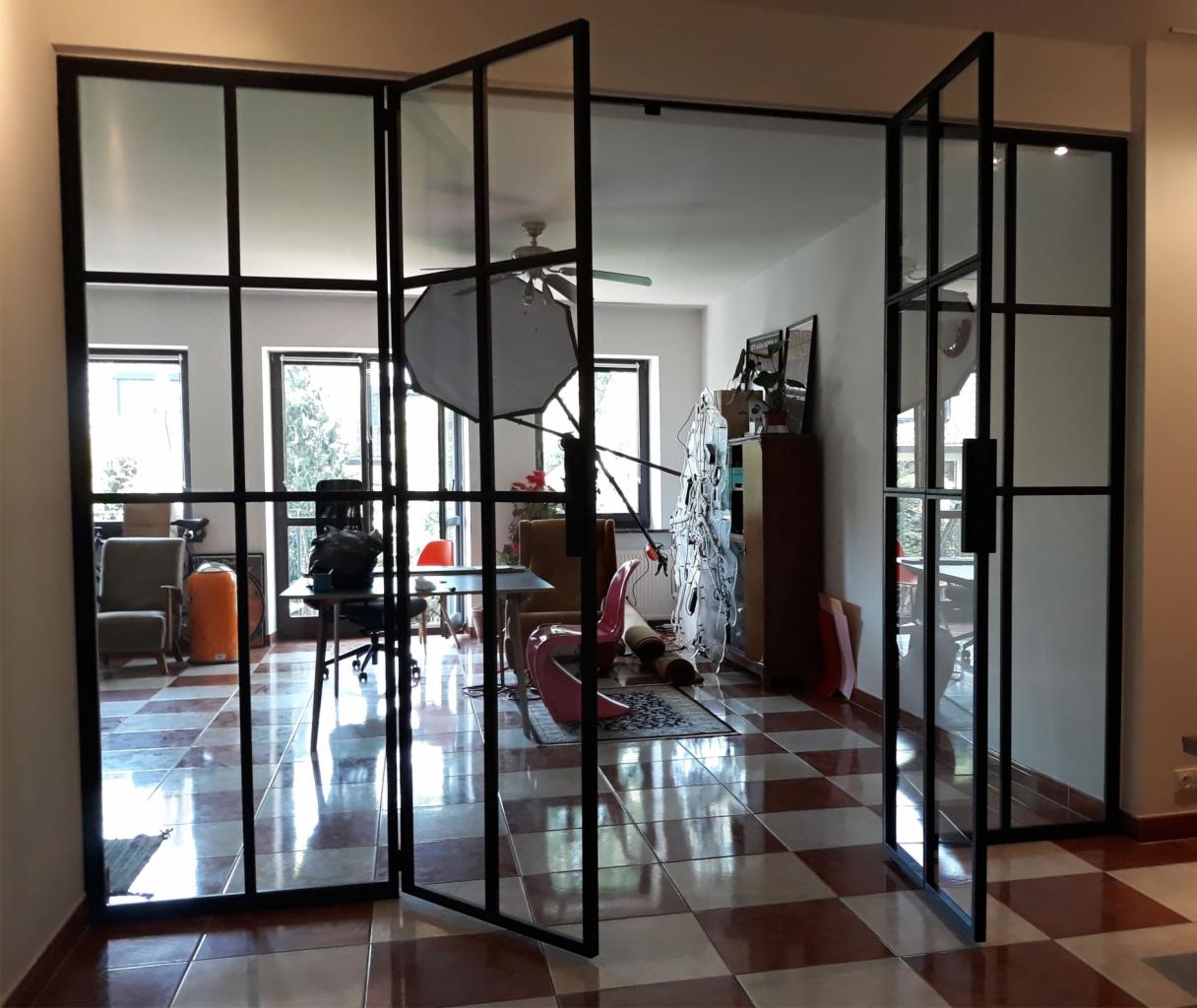 Otwierane, przeszklone drzwi wewnętrzne z czarnymi metalowymi szprosami, widok na pomieszczenie z szachownicową podłogą i wyposażeniem, częściowo widoczne oświetlenie studyjne.