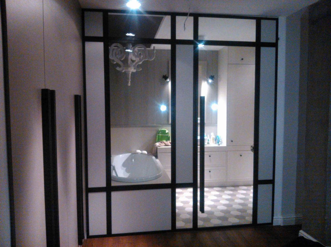 Przeszklona ścianka działowa z czarnymi profilami oddzielająca łazienkę od innego pomieszczenia, widoczna wanna wolnostojąca, białe meble łazienkowe i dekoracyjna lampa wisząca