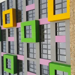 Wielopiętrowy budynek z szarą elewacją, różowymi akcentami i wystającymi, kolorowymi (żółtymi i zielonymi) kwadratowymi ramami okiennymi. Fragment ściany z cegły w trakcie budowy po prawej stronie.