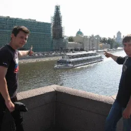 Dwaj mężczyźni wskazują na budynek w rusztowaniach i budynek w trakcie prac elewacyjnych na tle rzeki z przepływającym statkiem wycieczkowym.