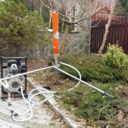 Wiertnica geologiczna z pomarańczowym cylindrem w ogrodzie, z widoczną pompą hydrauliczną i wężami, na tle kamiennego muru i drewnianego płotu.