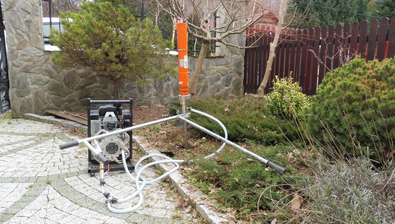 Wiertnica geologiczna z pomarańczowym cylindrem w ogrodzie, z widoczną pompą hydrauliczną i wężami, na tle kamiennego muru i drewnianego płotu.