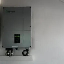 Szary falownik Growatt zamontowany na białej ścianie, widoczne przewody i dolna część czarnego elementu na ścianie po lewej stronie.