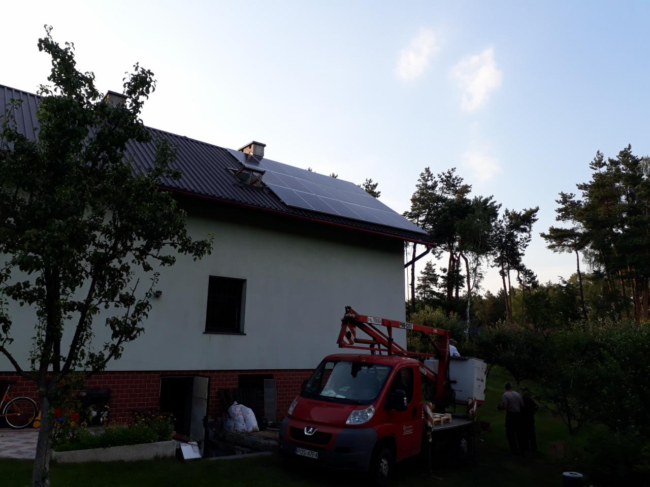 Montaż paneli słonecznych na dachu domu jednorodzinnego z użyciem podnośnika koszowego, widoczny pracownik w koszu i ekipa na ziemi.