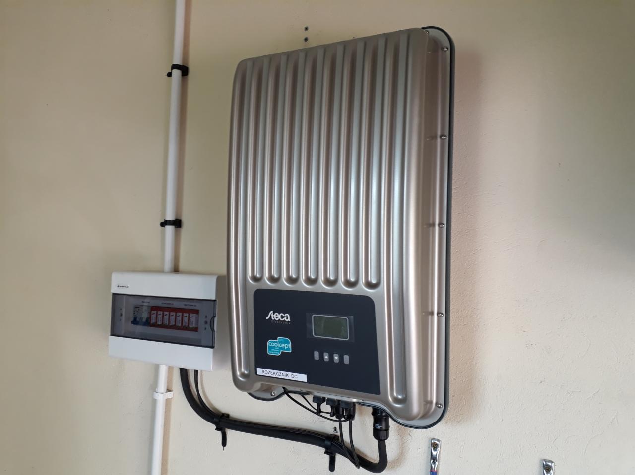 Szczegółowe ujęcie srebrnego inwertera marki Steca zamontowanego na beżowej ścianie, obok skrzynki elektrycznej z widocznymi bezpiecznikami.