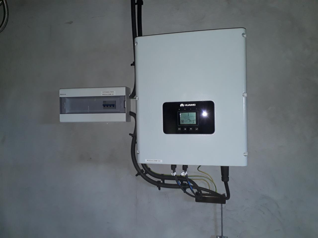 Biały inwerter Huawei z podłączonymi kablami i rozdzielnicą AC na szarej ścianie.