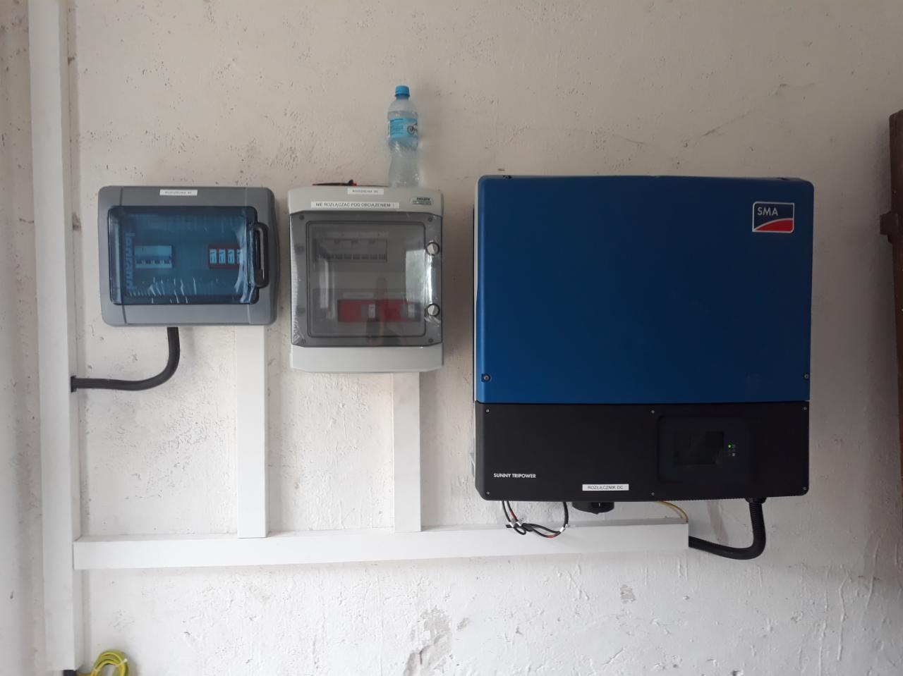 Instalacja fotowoltaiczna: niebieski inwerter SMA Sunny Tripower, dwie szare skrzynki elektryczne i butelka wody na jednej z nich, zamontowane na ścianie z białą listwą.