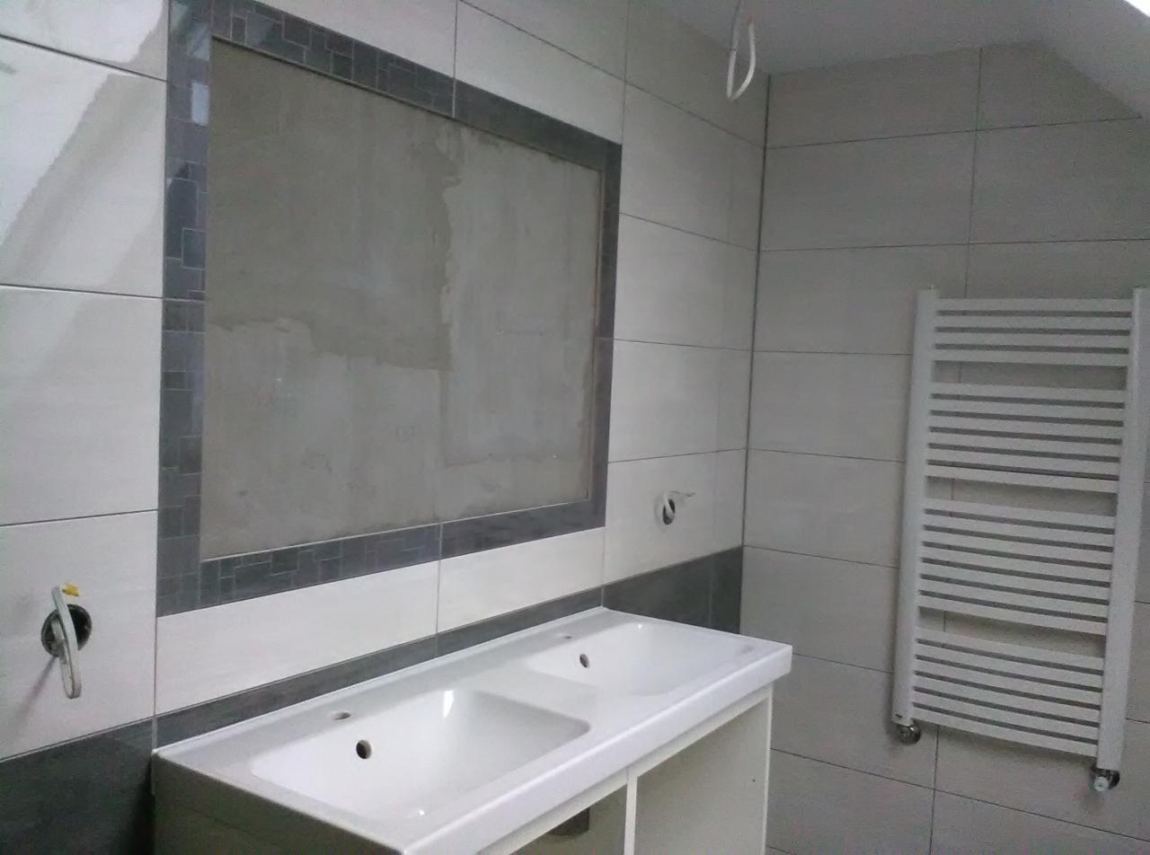 Łazienka w trakcie prac: białe płytki, umywalka podwójna z szafką, grzejnik drabinkowy na ścianie, wnęka na lustro z szarą mozaiką w trakcie wykańczania.