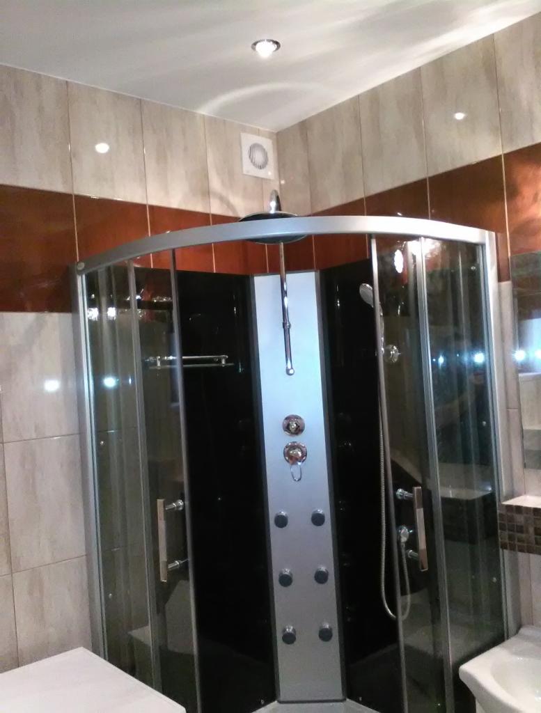 Nowoczesna kabina prysznicowa z hydromasażem, czarno-srebrna, w wykończonej łazience z beżowo-brązowymi płytkami i wentylatorem sufitowym.