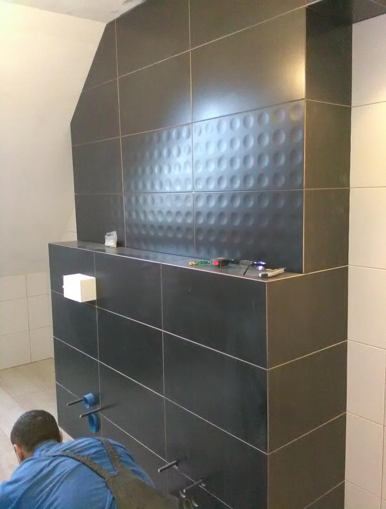 Częściowy widok łazienki w trakcie prac wykończeniowych; ciemne, prostokątne płytki z wypukłym wzorem ułożone na ścianie, widoczne narzędzia glazurnicze i fragment instalacji wodnej, pracownik...