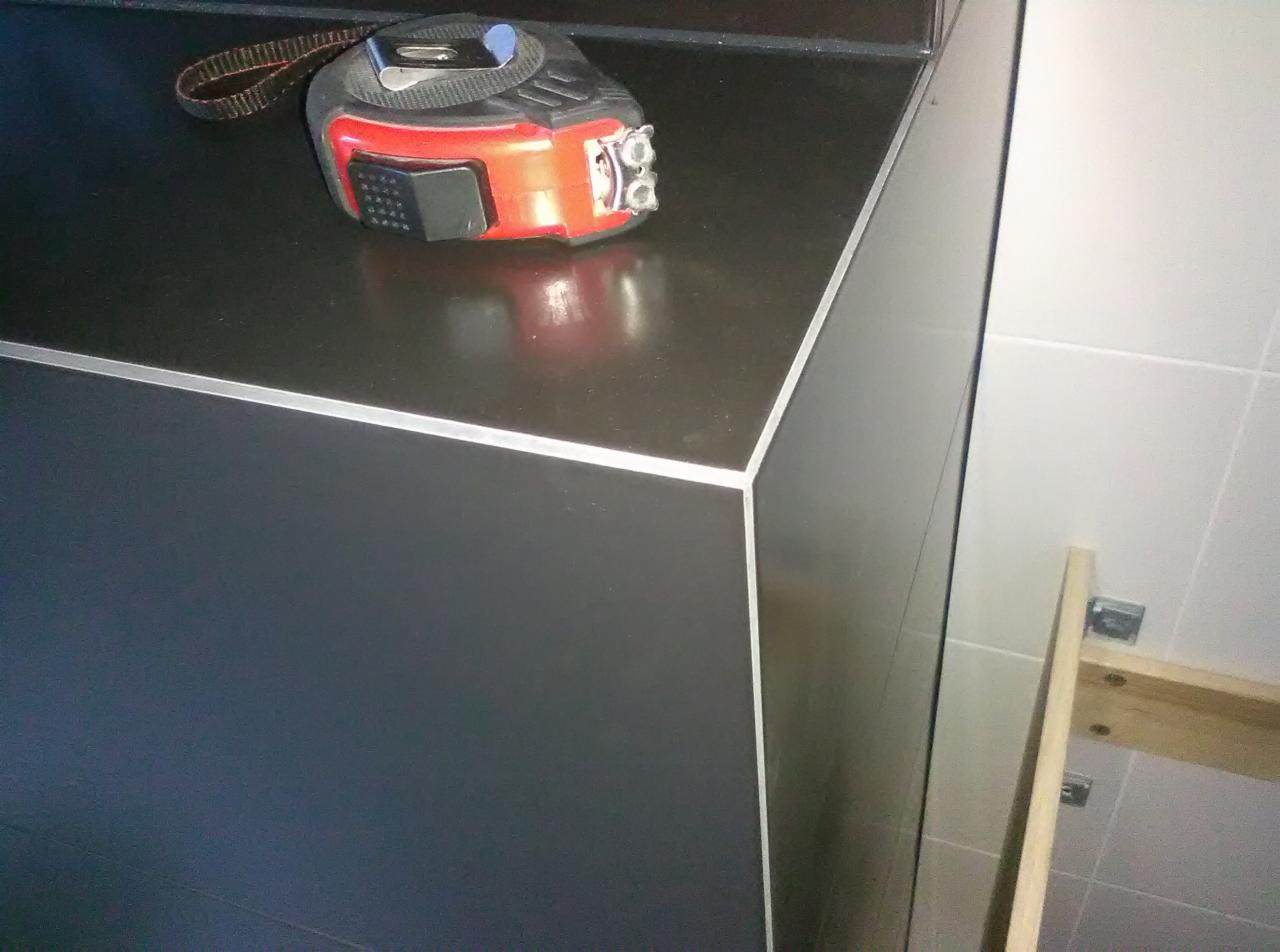 Czarny blat łazienkowy z aluminiowym wykończeniem krawędzi, na nim leży zwinięta miara zwijana, w tle fragment ściany z białych płytek i drewniana konstrukcja pod umywalkę.