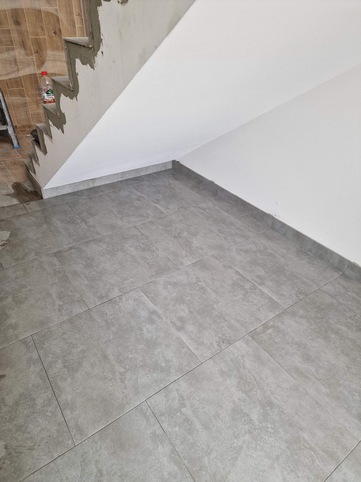Szare, prostokątne płytki podłogowe ułożone wzdłuż ściany i pod schodami w trakcie prac wykończeniowych, widoczna butelka z napojem i drabina w tle.