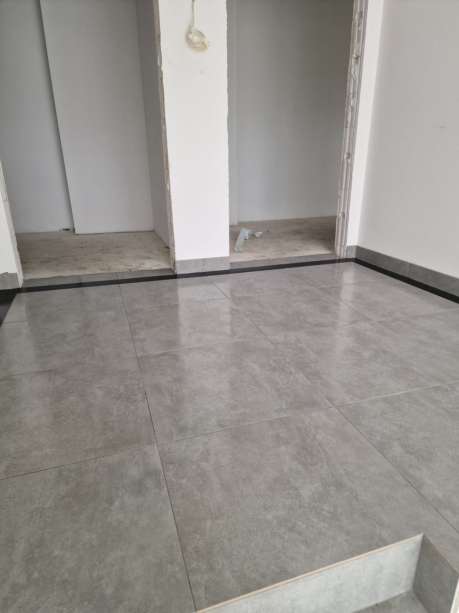 Ułożone szare płytki podłogowe z ciemnoszarą listwą przypodłogową w pomieszczeniu w trakcie remontu, widoczne surowe ściany i instalacja elektryczna.