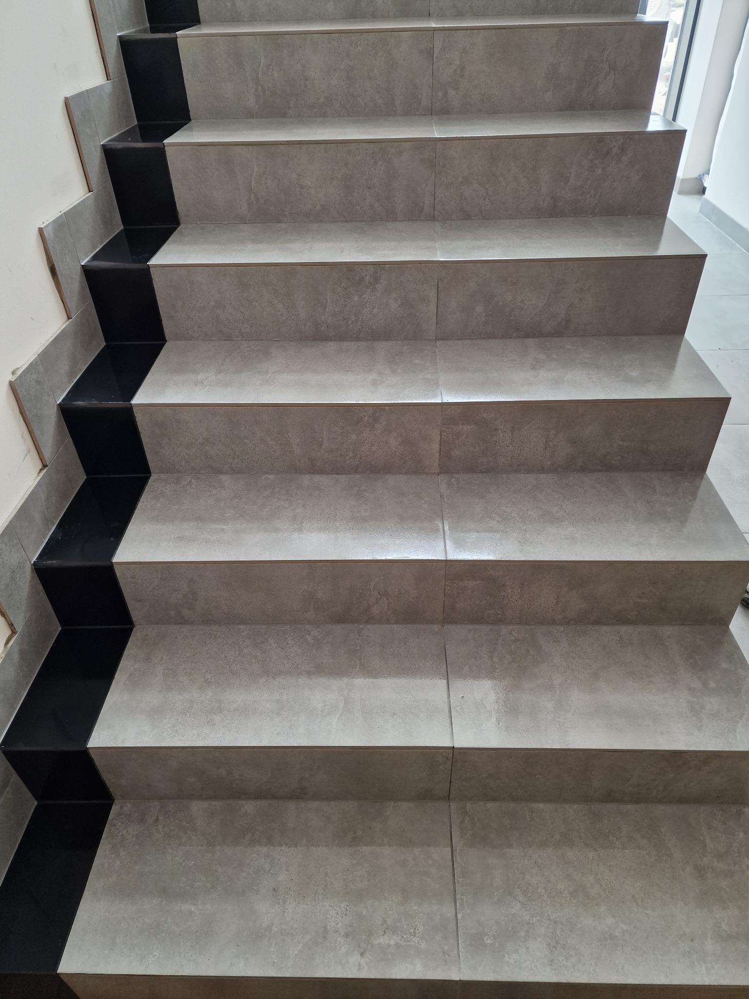Wykończone schody wewnętrzne z szarych płytek imitujących kamień, z czarnymi płytkami na bocznych krawędziach stopni.
