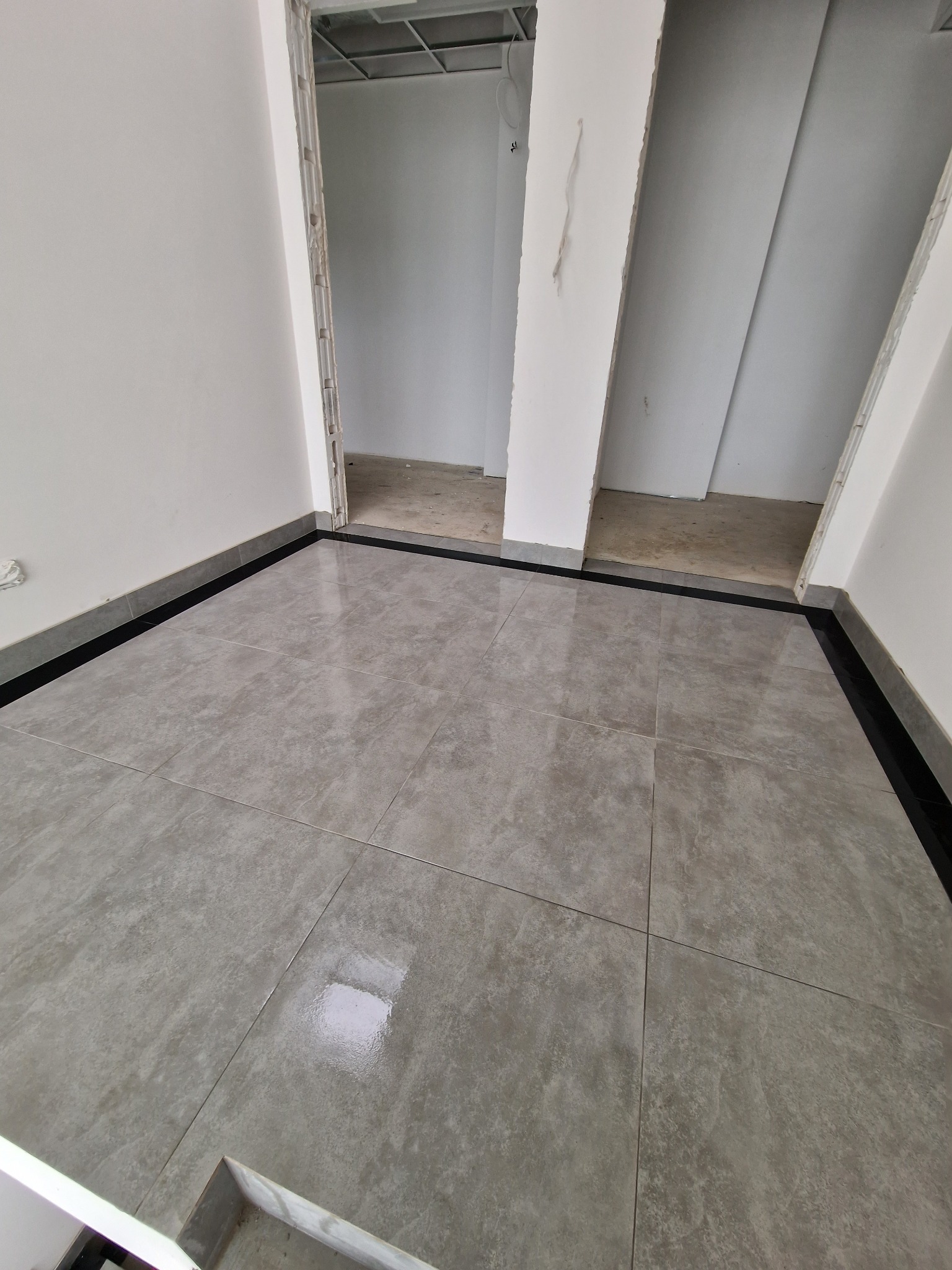 Szare płytki podłogowe z ciemną listwą przypodłogową w nowym, jeszcze niewykończonym pomieszczeniu. Widoczne surowe wnęki drzwiowe i fragment sufitu podwieszanego.