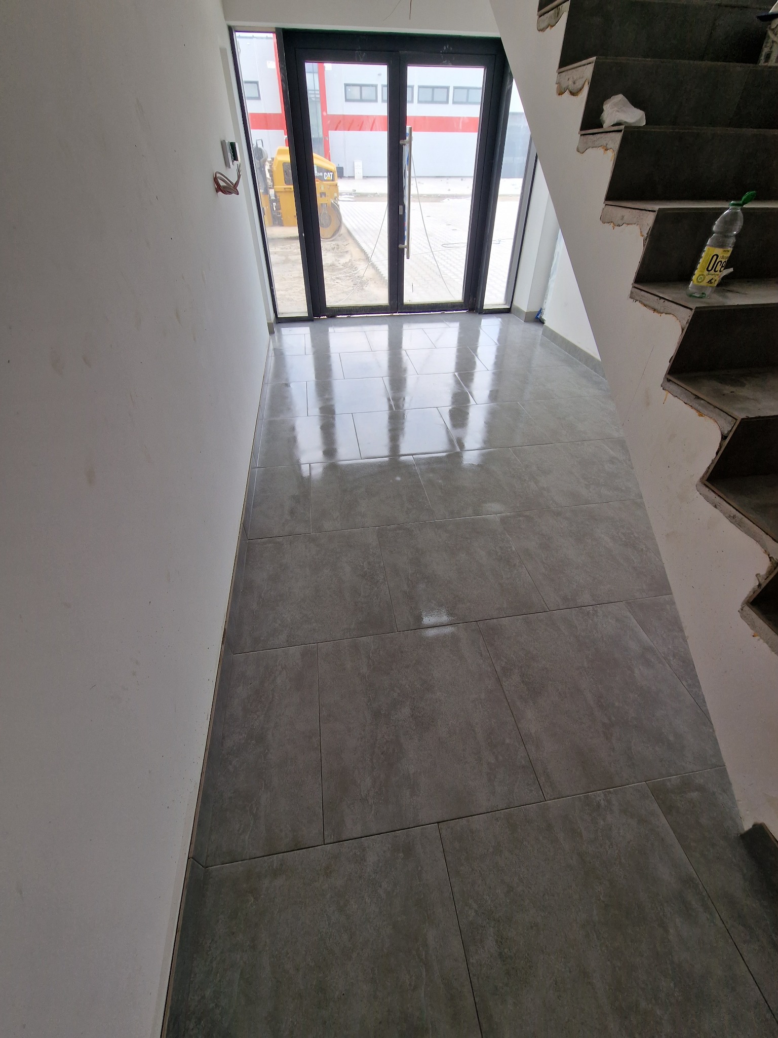 Ułożone szare płytki podłogowe w nowym budynku, widok na schody w stanie surowym i okno z widokiem na walec drogowy.