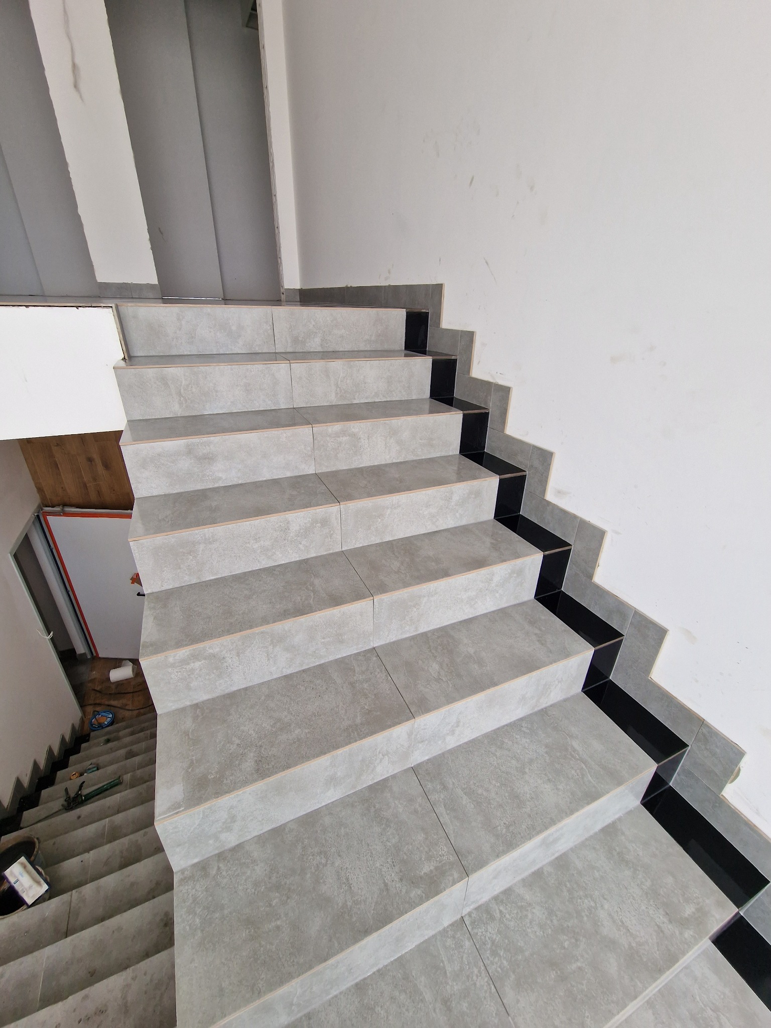 Schody wyłożone szarymi płytkami z czarnymi akcentami na krawędziach, widok z góry, narzędzia i akcesoria do układania płytek widoczne na dole schodów.