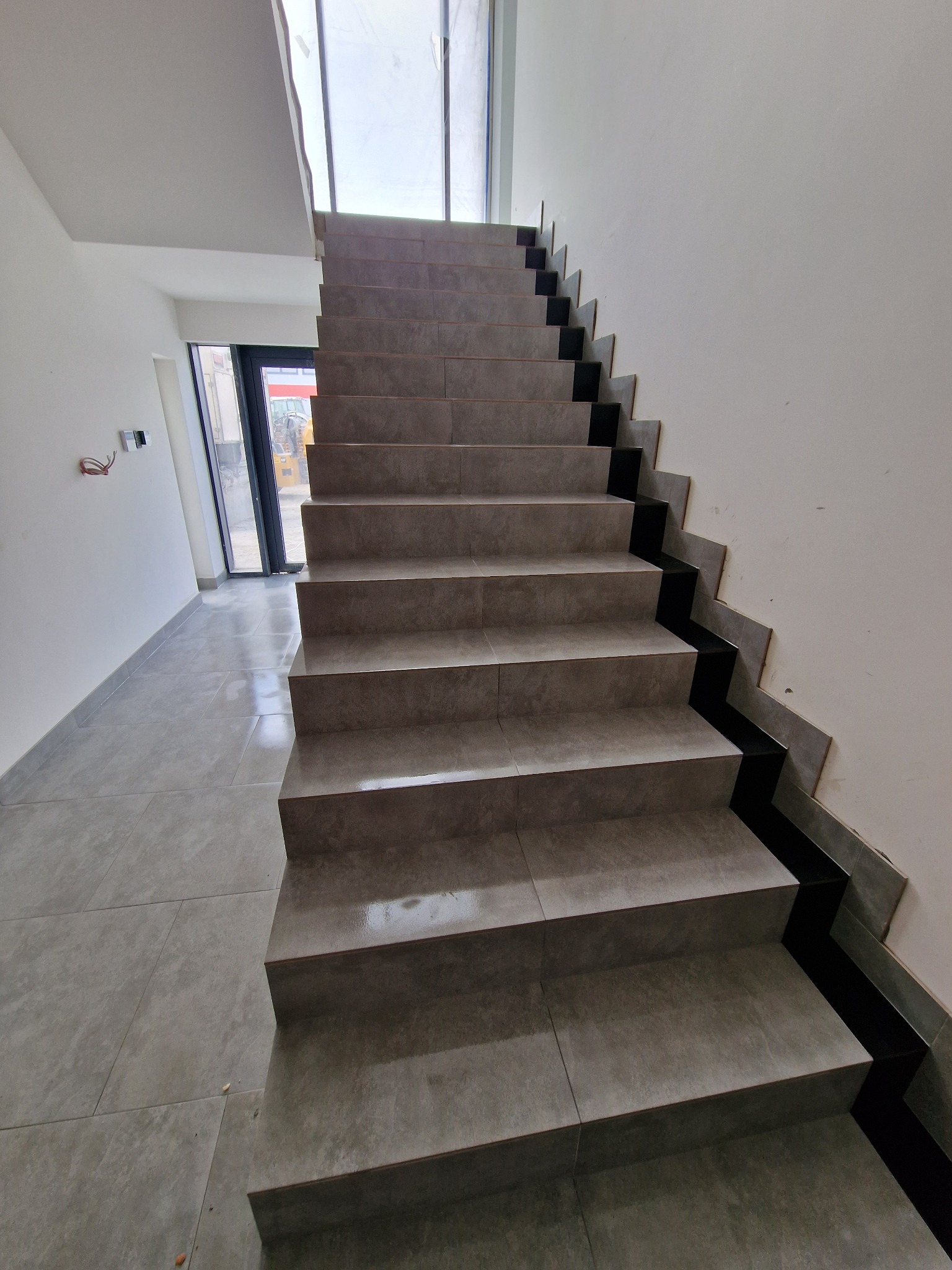 Wyłożone szarymi płytkami schody z czarnymi akcentami na krawędziach i podstopnicach, widok z dołu, w tle przeszklone drzwi z widocznym sprzętem budowlanym.