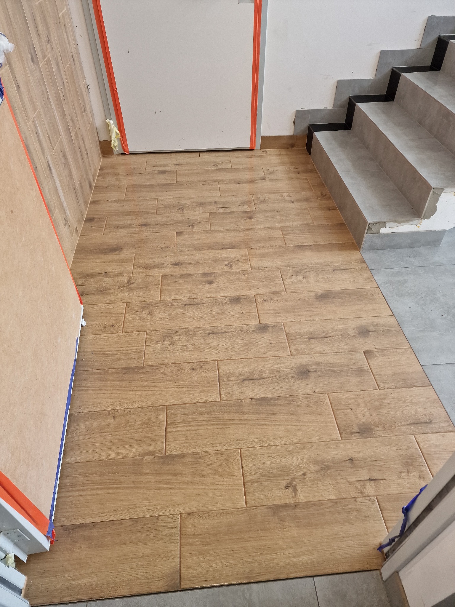 Ułożona podłoga z płytek imitujących drewno, widoczne schody wykończone szarymi płytkami, zabezpieczone folią malarską framugi drzwi.