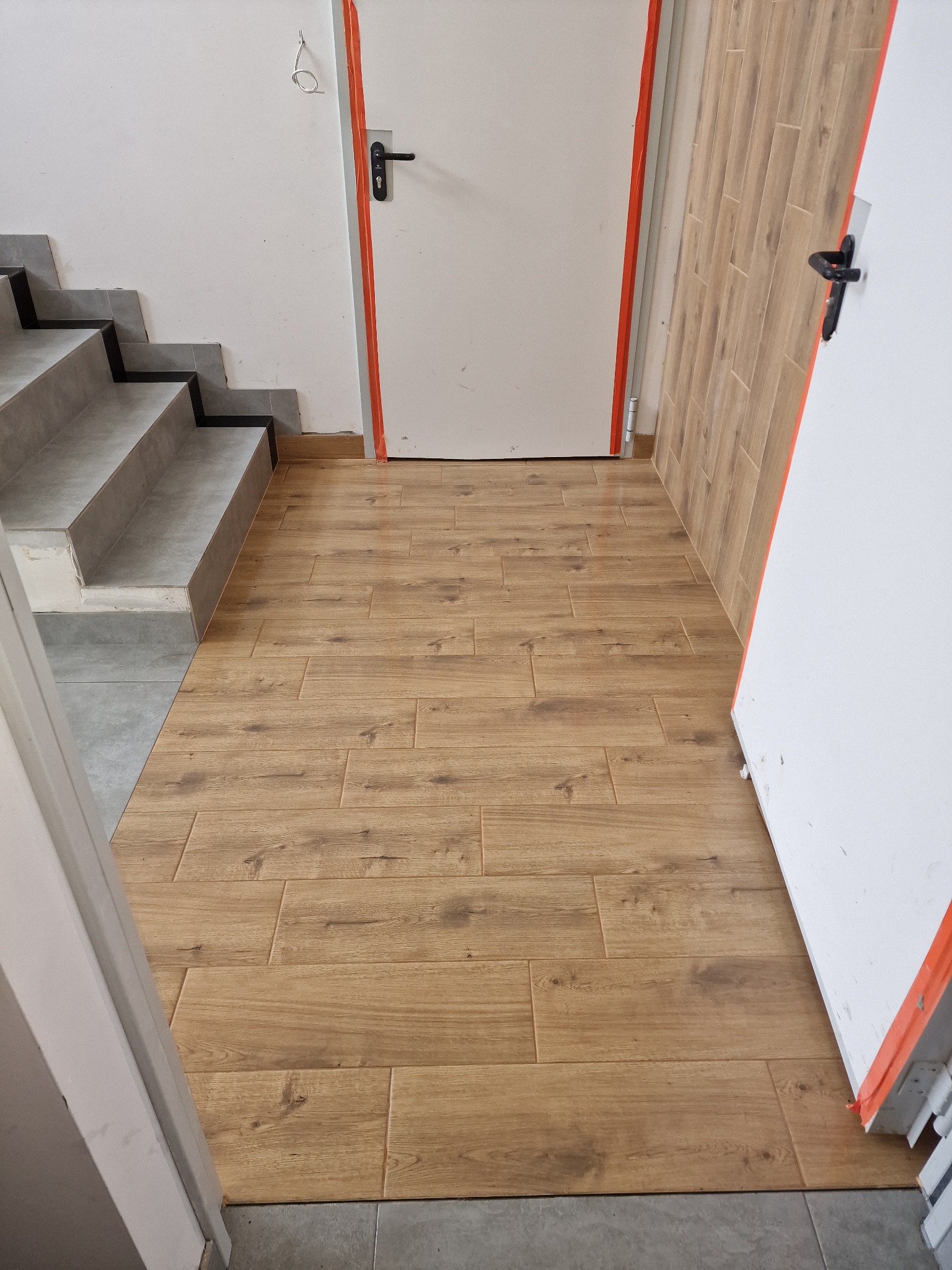 Ułożona podłoga z paneli imitujących drewno, łącząca się z szarymi płytkami schodów i fragmentem progu drzwiowego zabezpieczonego pomarańczową taśmą.