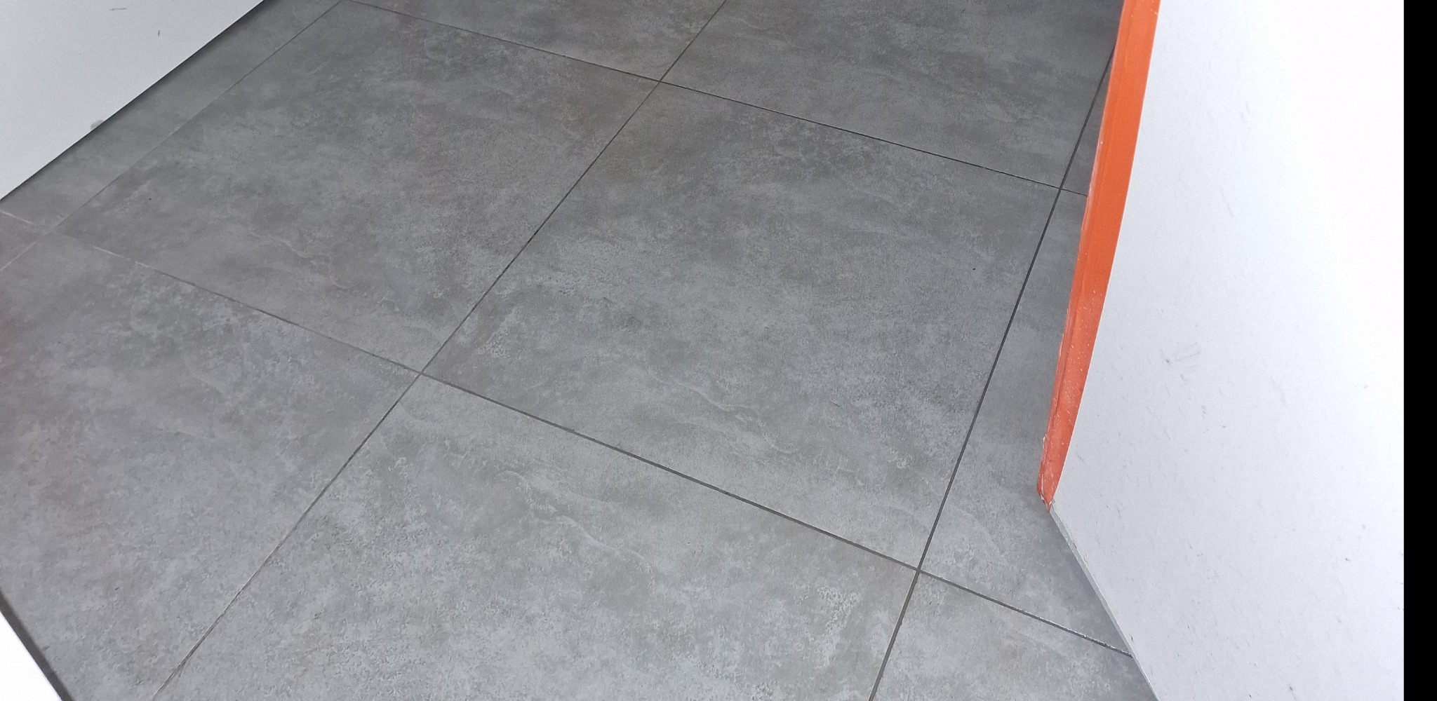Ułożona podłoga z szarych, matowych płytek gresowych imitujących kamień, z widoczną fugą i fragmentem białej ściany z pomarańczowym narożnikiem.
