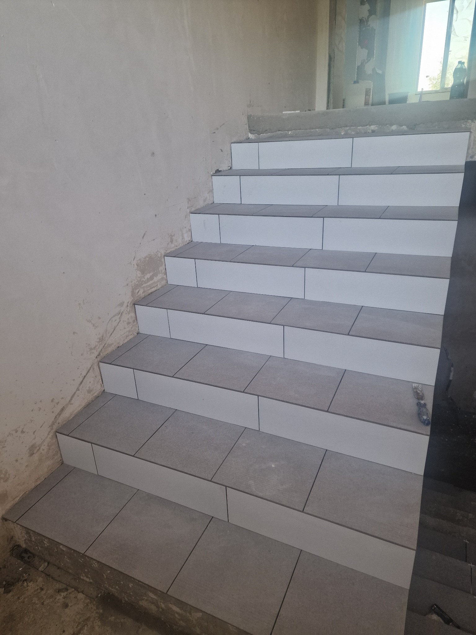 Wykończone płytkami schody w surowym stanie; szare płytki na stopniach i białe na podstopnicach, widoczne niedoskonałości ścian i brak listew przypodłogowych.