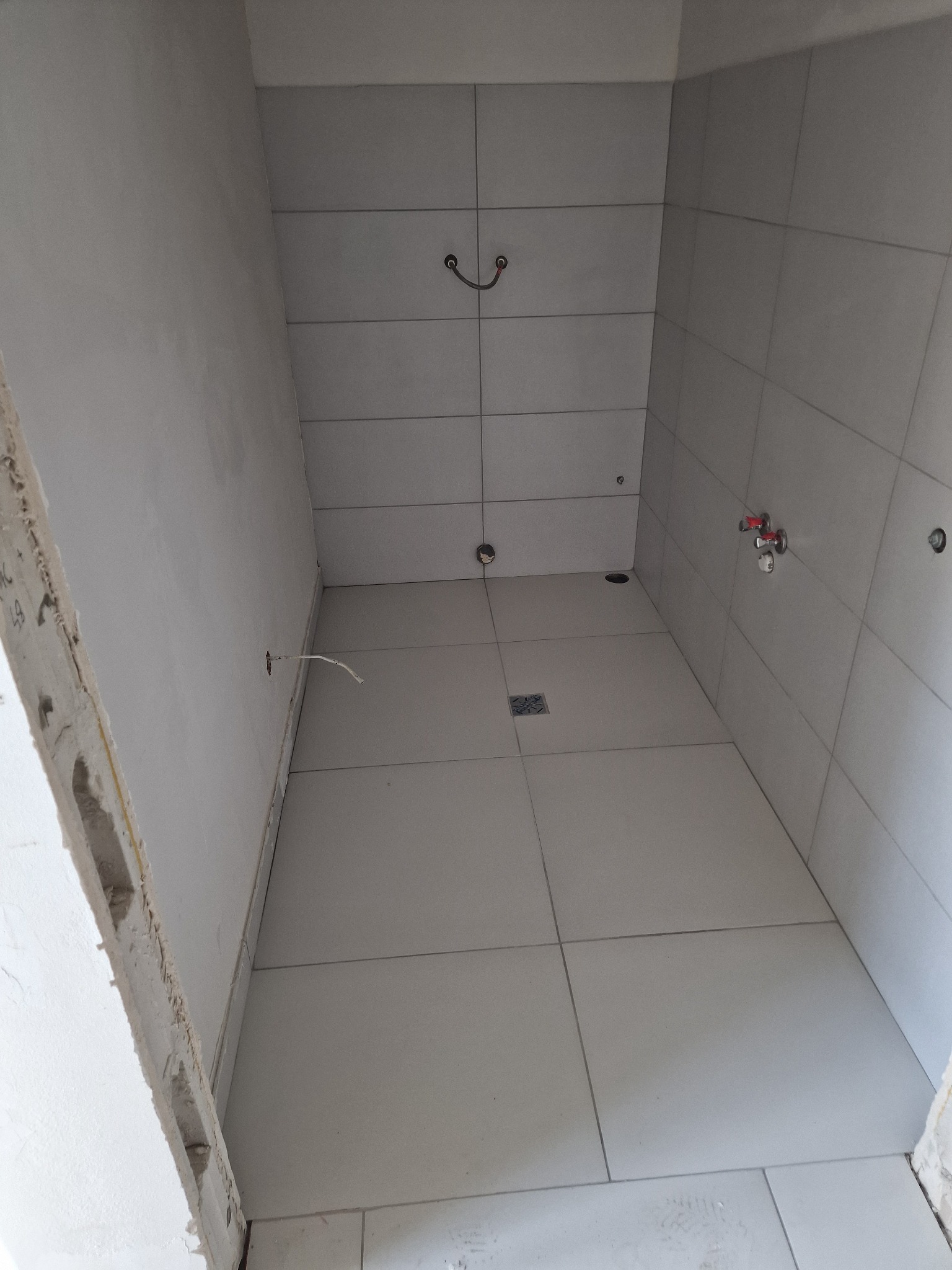 Płytki ceramiczne w jasnym odcieniu szarości, ułożone w kabinie prysznicowej z widocznym odpływem liniowym i wystającymi rurami instalacji wodnej, ściany przygotowane do dalszych prac wykończeniowych.