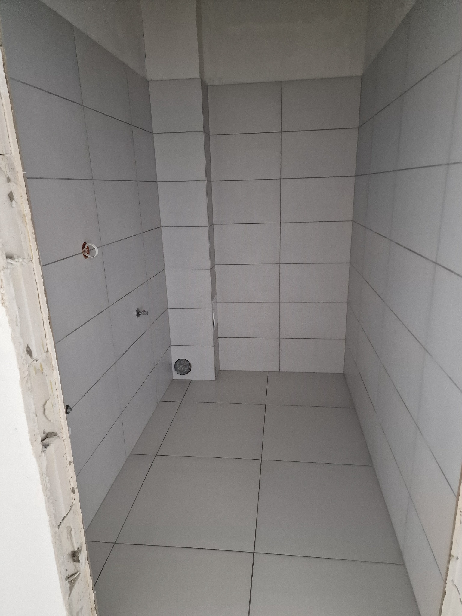 Wnętrze wykończonej łazienki z jasnymi, kwadratowymi płytkami ceramicznymi na ścianach i podłodze, widoczne fragmenty instalacji hydraulicznej i elektrycznej, surowa ościeżnica drzwiowa po lewej...