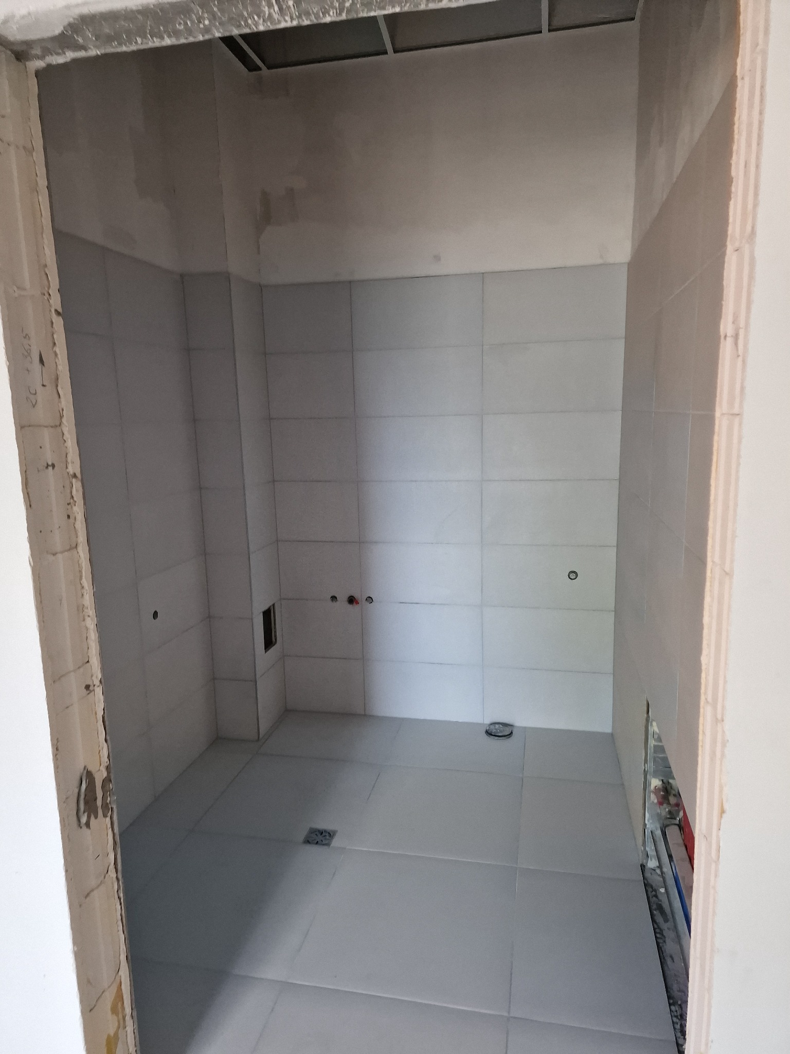 Wnętrze łazienki w trakcie prac wykończeniowych: ułożone jasne płytki na ścianach i podłodze, widoczne punkty instalacji i odpływ liniowy; surowy ościeżnica drzwiowa z resztkami farby.